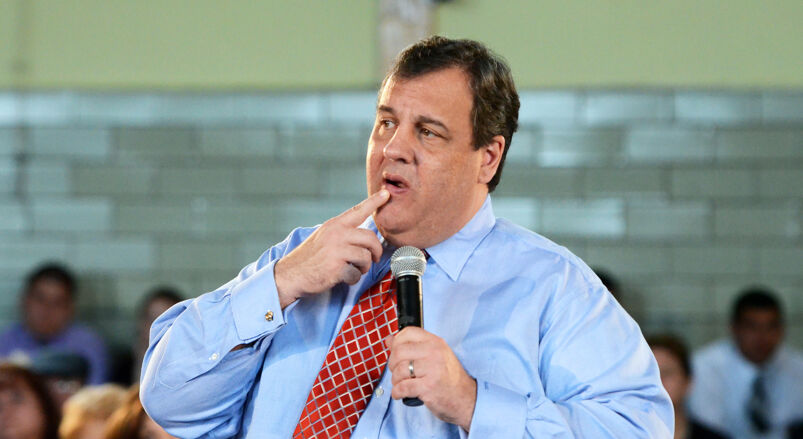 Chris Christie in a blue shirt and red striped tie holding a microphone