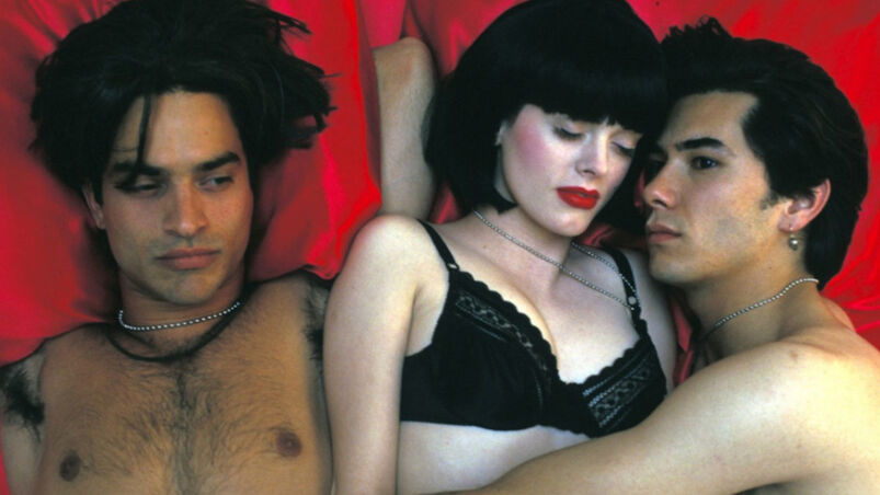 Three shirtless people lay next to each other in bed on bright-red sheets. The man on the right has his arm around the woman in the middle, who is wearing a black bra and has a bob haircut.