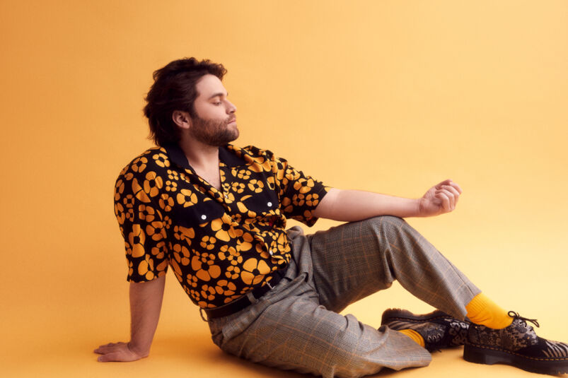 Actor Jake Borelli wears a floral=printed button-up shirt and grey slacks while sitting in front of a yellow backdrop