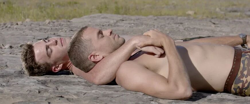 Two shirtless soldiers lay on the dirt in one another's arms.