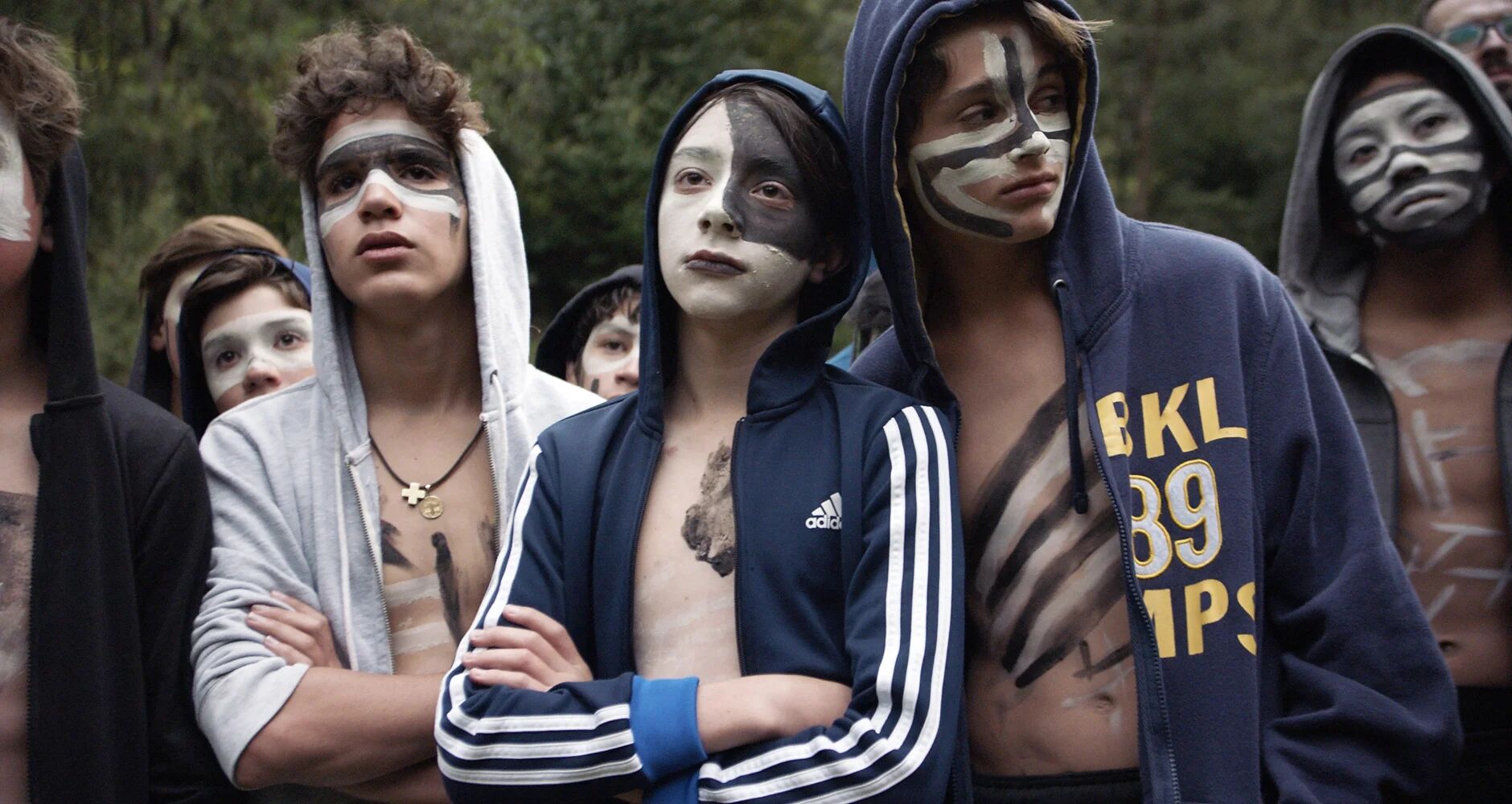 A group of young men wears hoodies and striking black-and-white face paint with trees behind them