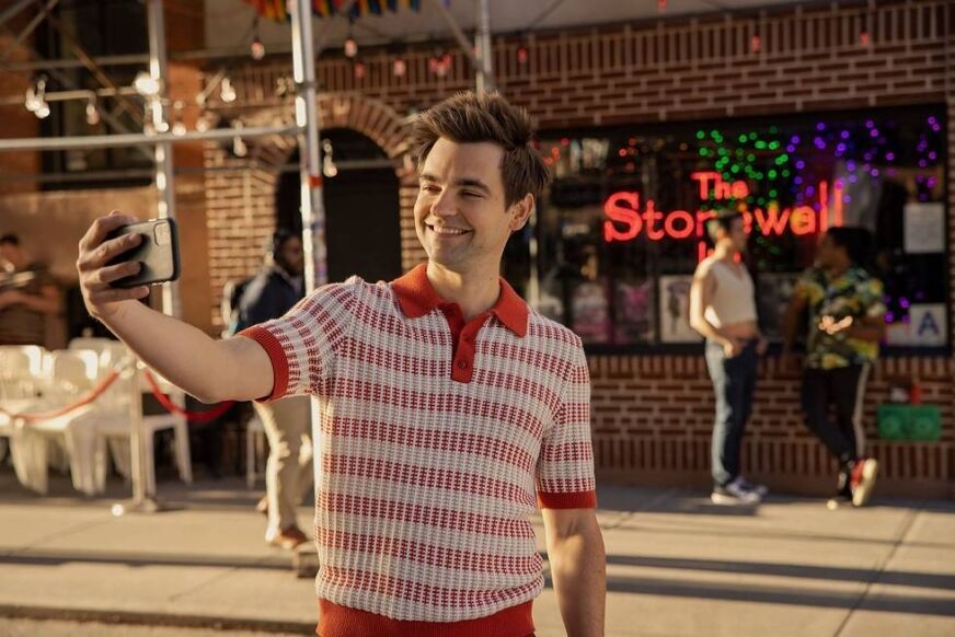 Drew Tarver wears a red and white knit polo, posing to take a selfie in front of The Stonewall Inn