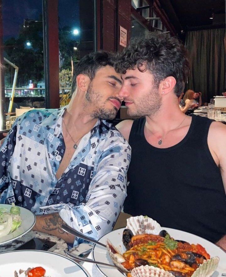 Actors Santi Talledo and Toni Gelabert embrace while sitting side-by-side at a restaurant