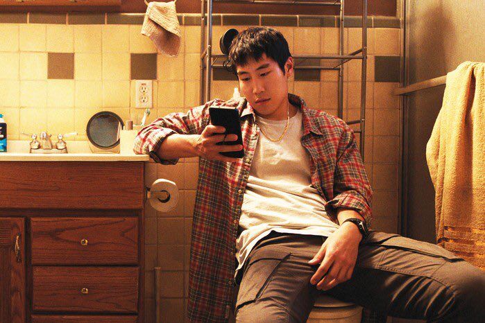 Young Mazino in 'Beef,' looking at his phone while sitting and wearing red flannel over a white tee.