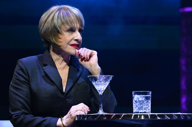 Patti LuPone wears a black shirt and has a martini while on stage