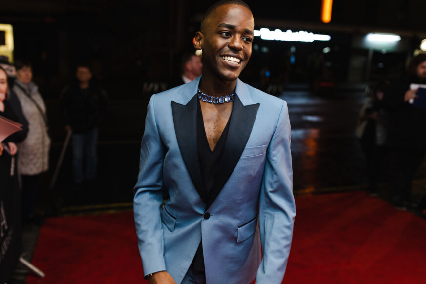 Actor Ncuti Gatwa poses and smiles on a red carpet, wearing a light blue jacket with black lapels and no shirt underneath.