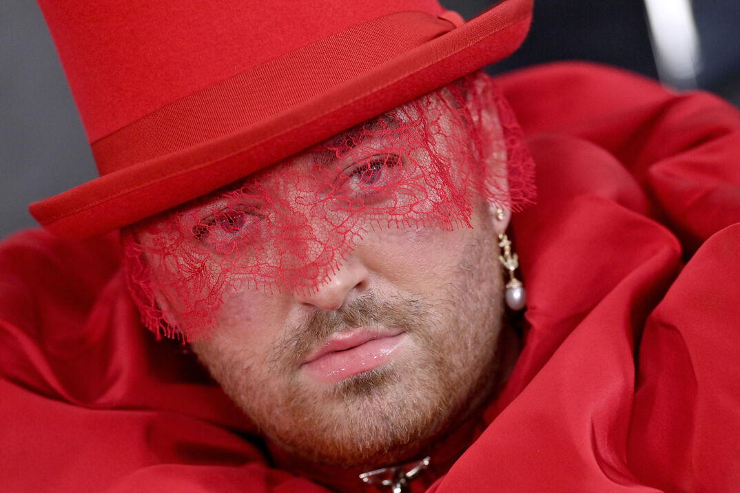 Sam Smith in a red dress, pearl earrings, and a red lace mask.