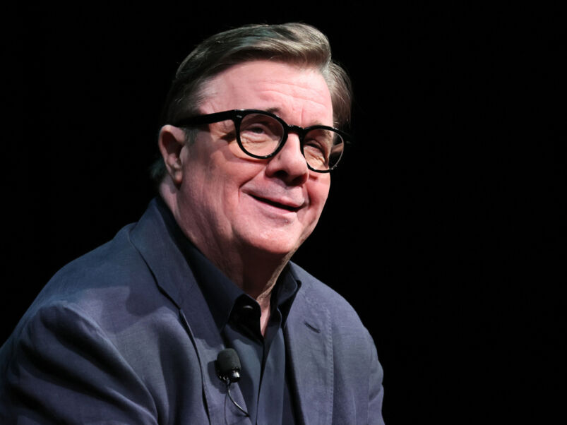 Nathan Lane wearing dark-rimmed glasses and a navy blue suit in front of a black background.