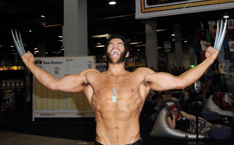 A buff shirtless man cosplays as Wolverine wearing dog tags on a chain and fake claws
