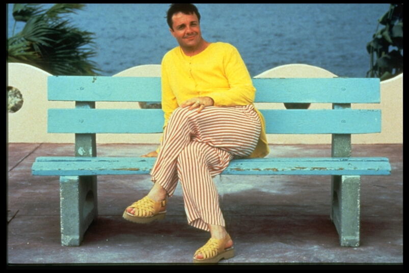 Nathan Lane wears an all-yellow outfit while sitting on a blue park bench in front of the ocean in 'The Birdcage'