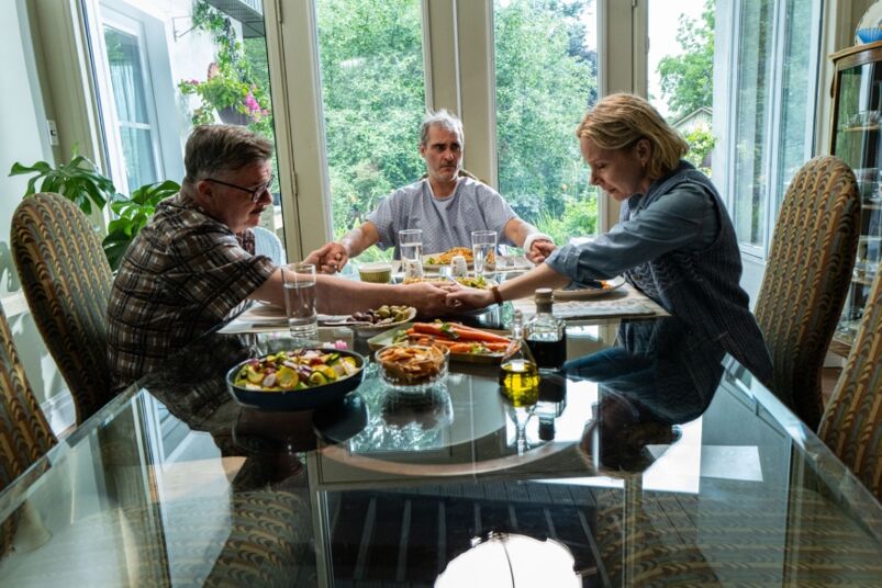 Nathan Lane, Joaquin Phoenix, and Amy Ryan hold hands around a dinner table full of food in 'Beau is Afraid'