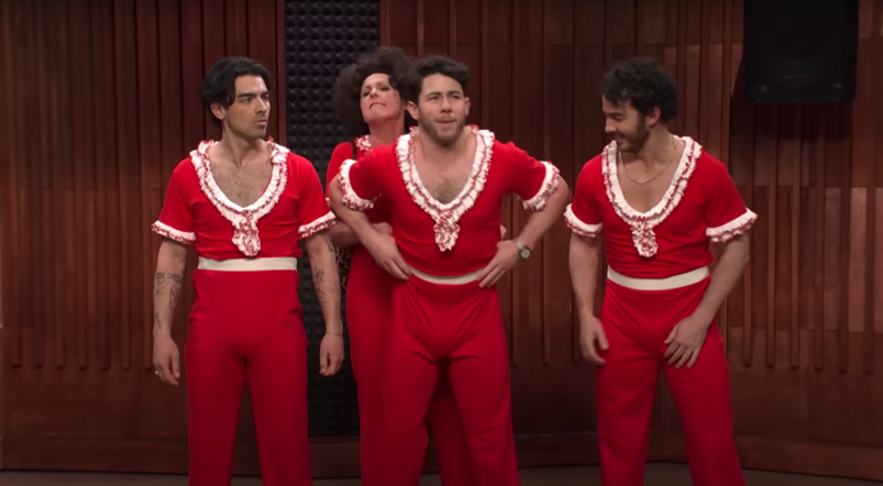 The Jonas Brothers stand in a row in the middle of a dance studio, each wearing revealing red otufits, with Molly Shannon behind them.