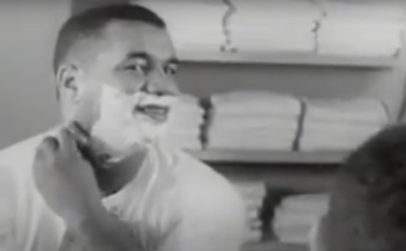 Black man with short hair shaving himself with shaving cream on his face and wearing a white t-shirt.