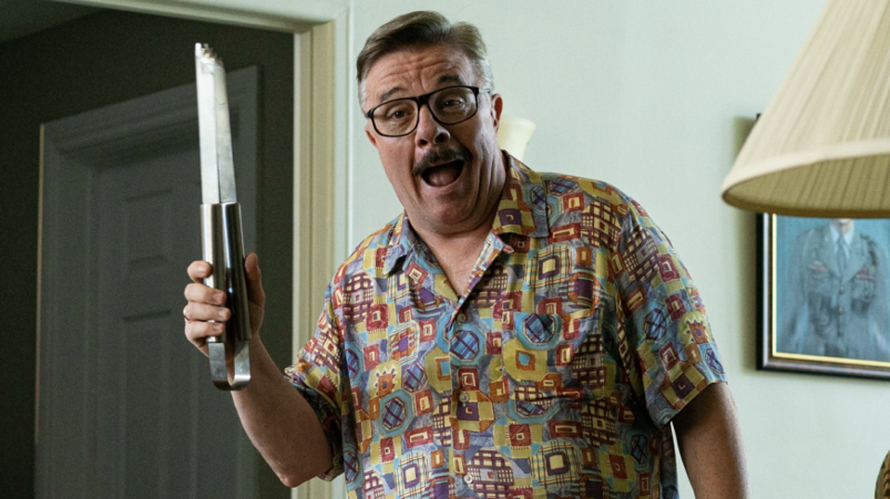 Nathan Lane sports. mustache and wears a loud, patterned shirt while holding grill tongs.
