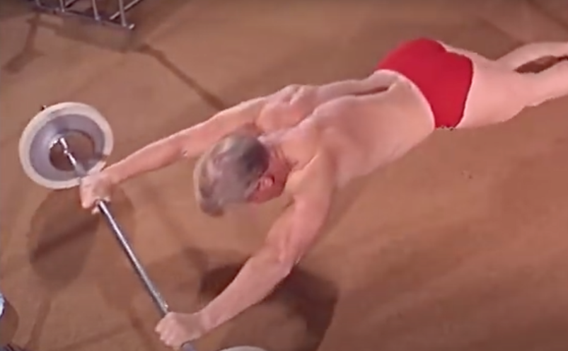 Shirtless man with blonde hair wearing a red speedo and stretching himself out.