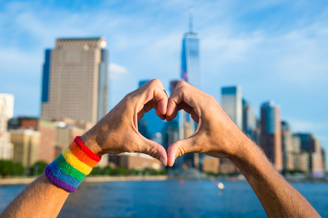 Hands wearing gay pride rainbow sweatband making a handheart symbol in front of city skyline in New York
