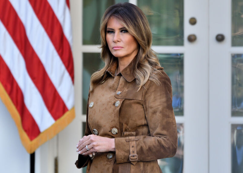Melania Trump in a brown coat standing in front of an American flag