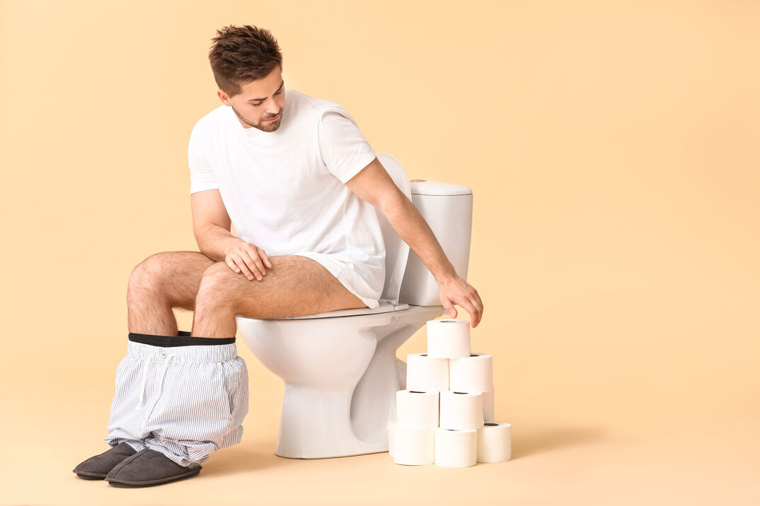 A man sitting on a toilet with his pants down and a pyramid of toilet paper stacked next to him