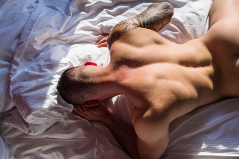 a naked man laying face down on a bed with his butt in the air but cropped off screen