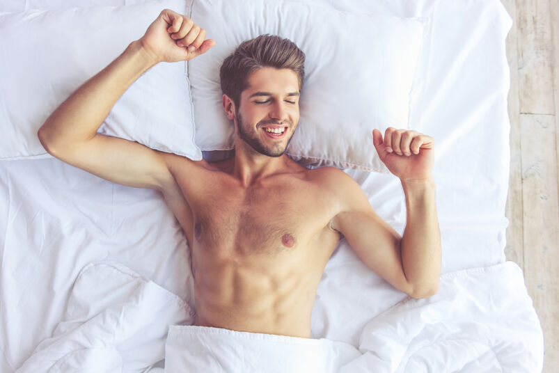 A smiling, shirtless man laying in bed with white sheets
