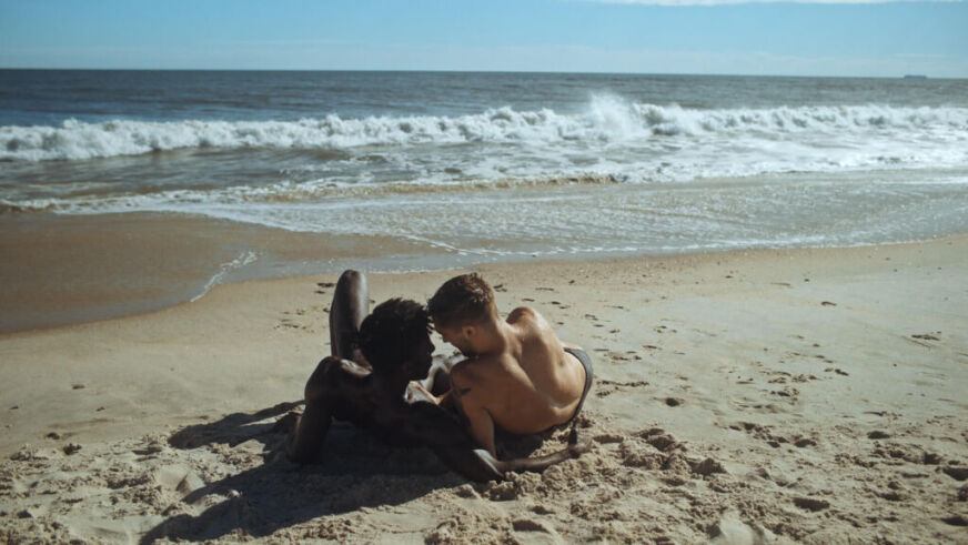 Two muscled men lay shirtless on a beach, staring into one another's eyes, with the ocean in front of them.