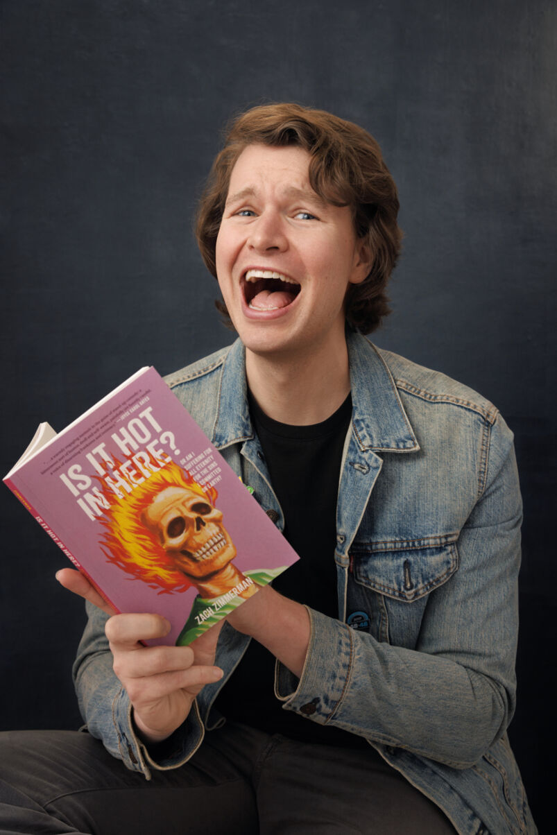 Comedian Zach Zimmerman wears a denim jacket and laughs while reading his book 'Is It Hot In Here?'