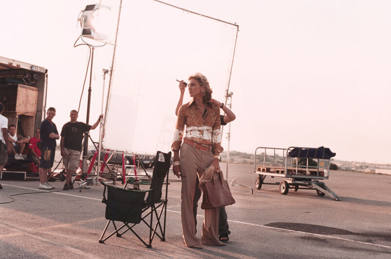 Madonna, wearing a golden button up, pressed slacks heels, and carrying a purse, glamorously poses while getting her makeup touched up behind the scenes of 'Swept Away.'