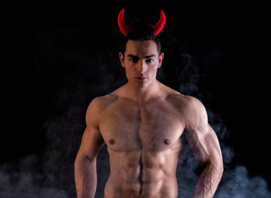 A muscled shirtless man wears red devil horns and stands in front of a black background