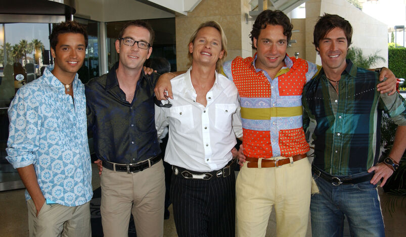 'Queer Eye for the Straight Guy' stars Jai Rodriguez, Ted Allen, Carson Kressley, Thom Filicia, and Kyan Douglas stand smiling with their arms around each other in front of a hotel.