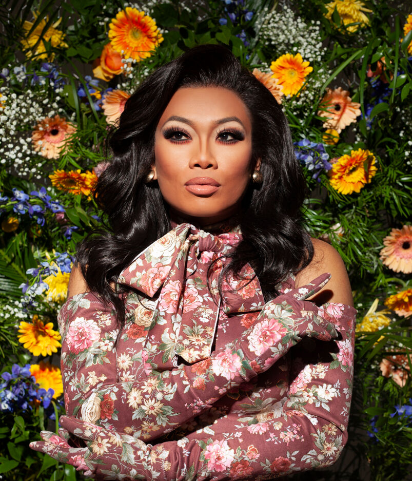 Drag queen Jujubee wears floral print dress and black wig, posing in front of wall of flowers