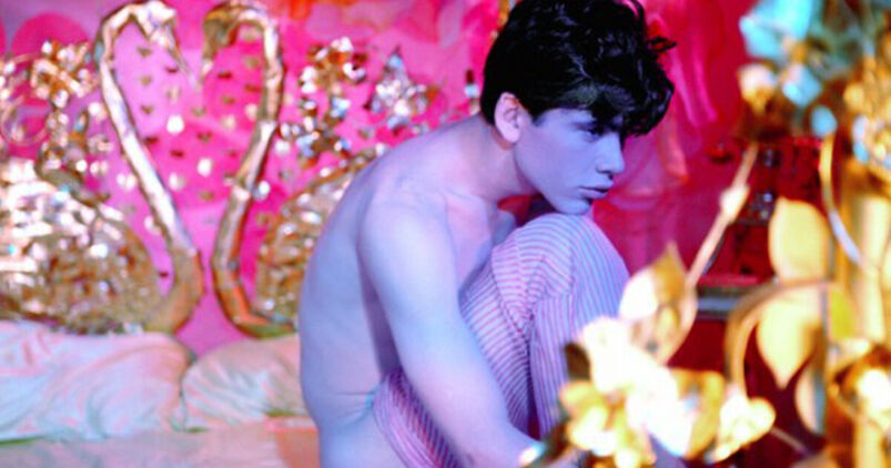 A still from 'Pink Narcissus' featuring a call-boy in underwear sitting on a bed with a pink wall behind him.