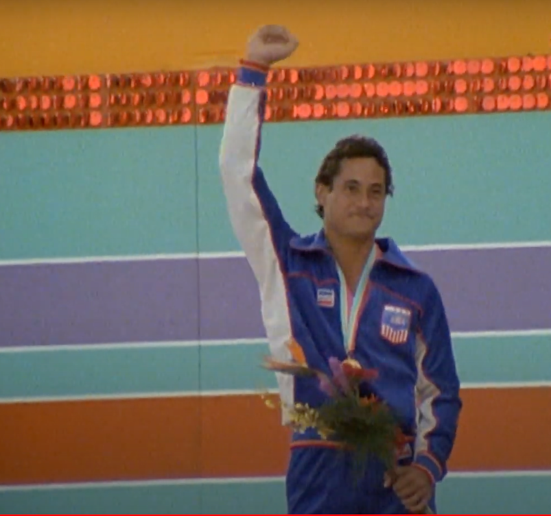 Greg Louganis posing in his Olympic uniform with a medal around his neck and flowers.