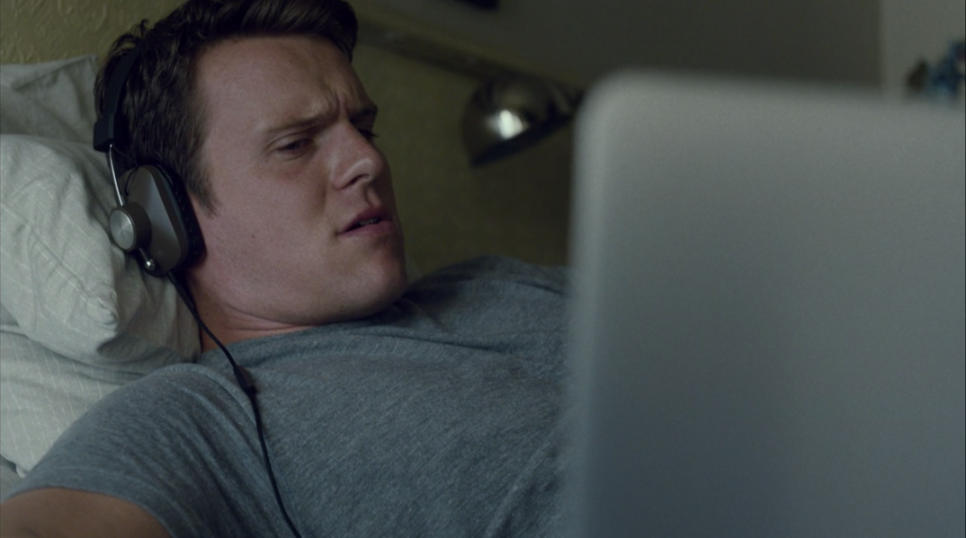 Jonathan Groff wearing headphones laying in bed looking at a laptop screen.