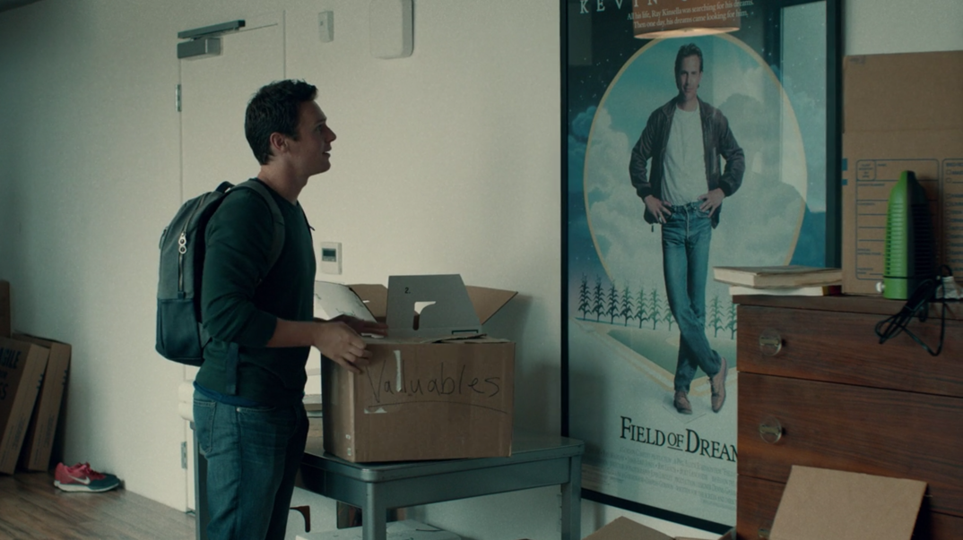 Jonathan Groff, holding a moving box, examines a 'Field of Dreams' poster hanging on an apartment wall.