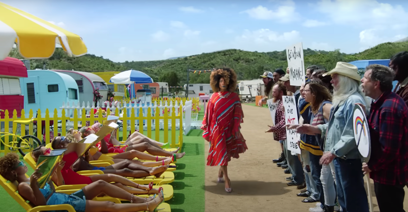 Billy Porter, wearing a red dress, walks in front of a crowd of angry anti-LGBTQ+ protestors while Taylor Swift and her friends tan in lounge chairs in a scene from the "You Need to Calm Down" music video.