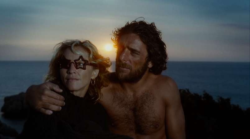 Madonna, wearing broken star-shaped sunglasses, looks on while a shirtless Adriano Giannini wraps his arm around her in front of a setting sun in a scene from 'Swept Away.'