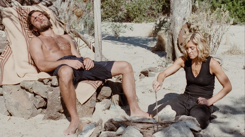 A shirtless Adriano Giannini relaxes on a throne made of rocks on a deserted island while Madonna tries to cook a fish with a stick in a rock pit in a scene from 'Swept Away.'
