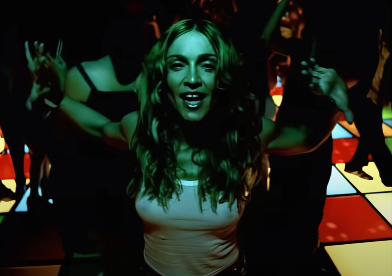 Madonna dances on a colorful dance floor wearing a white tank top in a scene from the "Ray of Light" music viideo.