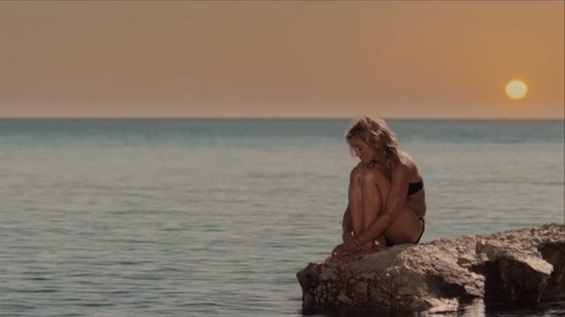 Madonna, in a black bikini, sadly sits on a rock in the ocean looking at her toes as the sun sets in a scene from 'Swept Away.'