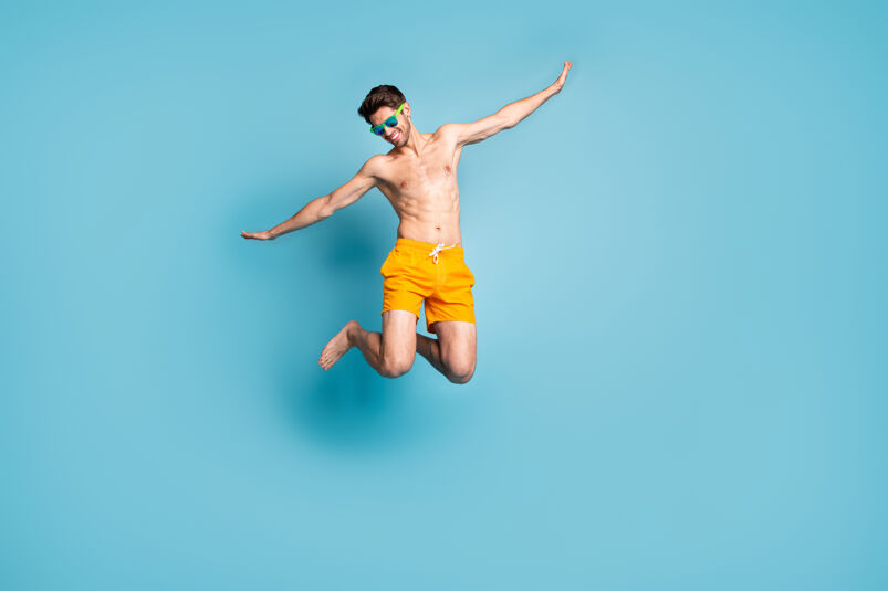 Shirtless guy wearing sunglasses leaping in the hair with an orange swimsuit.