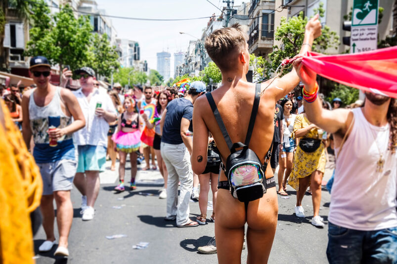 A man wearing a backpack and a revealing jockstrap is pictured from the back, waving at attendees at an outdoor Pride festival.