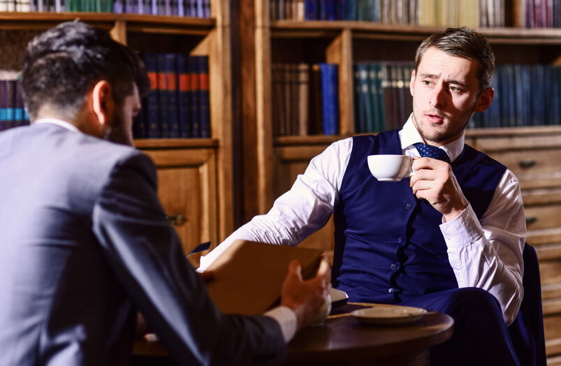 A dapper man drinking tea across from a man in a suit. 
