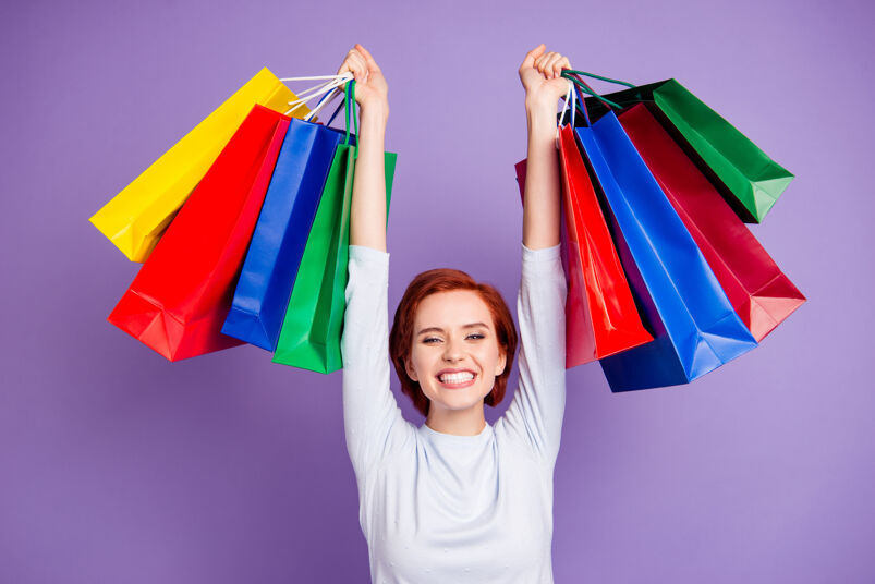 A smiling woman holds her hands above her head, carrying a large assortment of rainbow colored shopping bags.