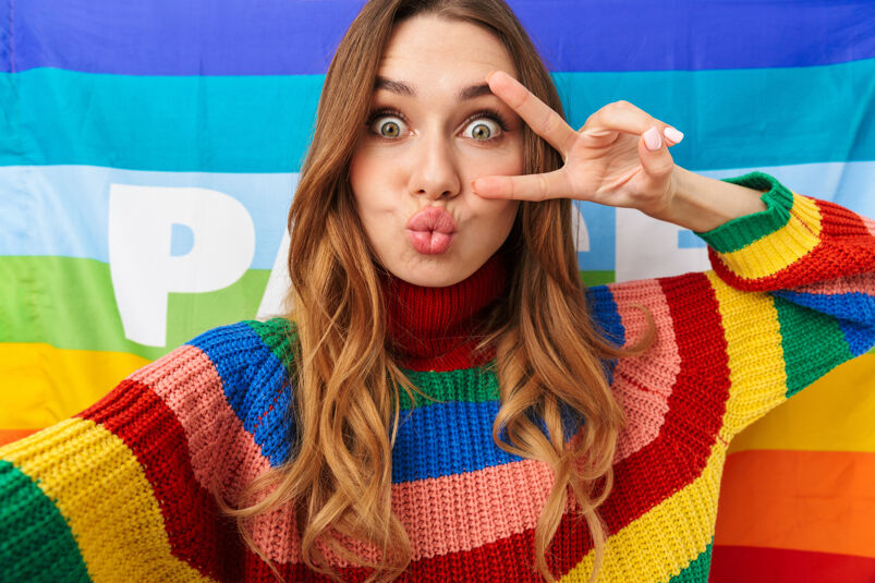 A girl in a knitted rainbow sweater, posing in front of a Pride flag, takes a selfie while holding up a peace sign.