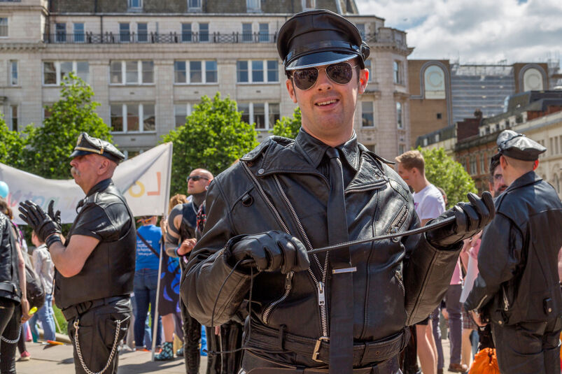 A man dressed in full leather gear smiles and holds a whip at an outdoor Pride festival. 