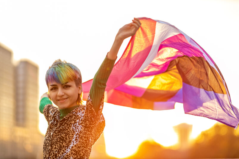 Nonbinary person with rainbow hair and leopard shirt waving the Gender Fluid Pride Flag.