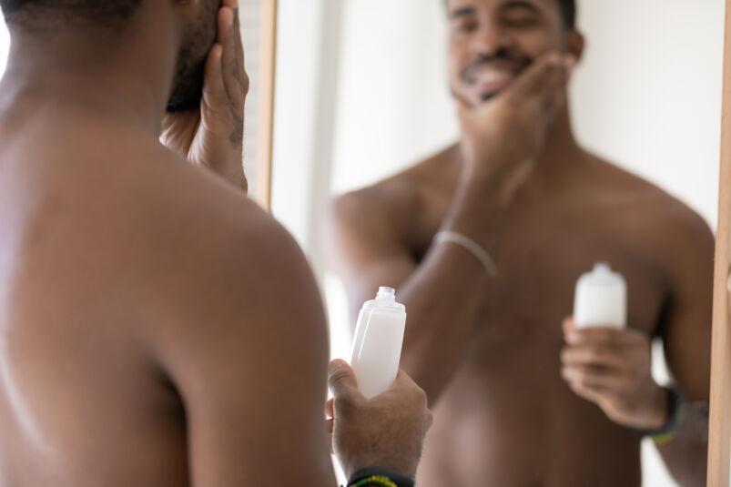 Man looking at himself in the mirror with a smile and applying after shave. 
