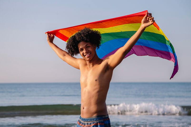 A black man holding the Pride Flag on the beach. 
