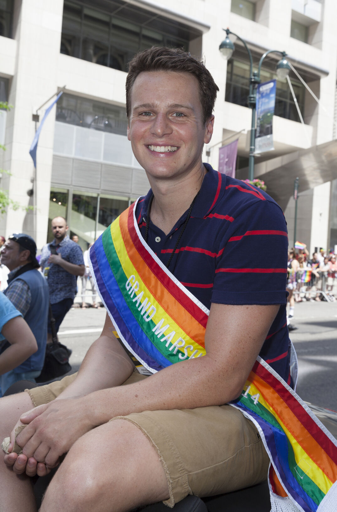 Jonathan Groff smiles while wearing a rainbow sash reading "Grand Marshal" outside in New York City.
