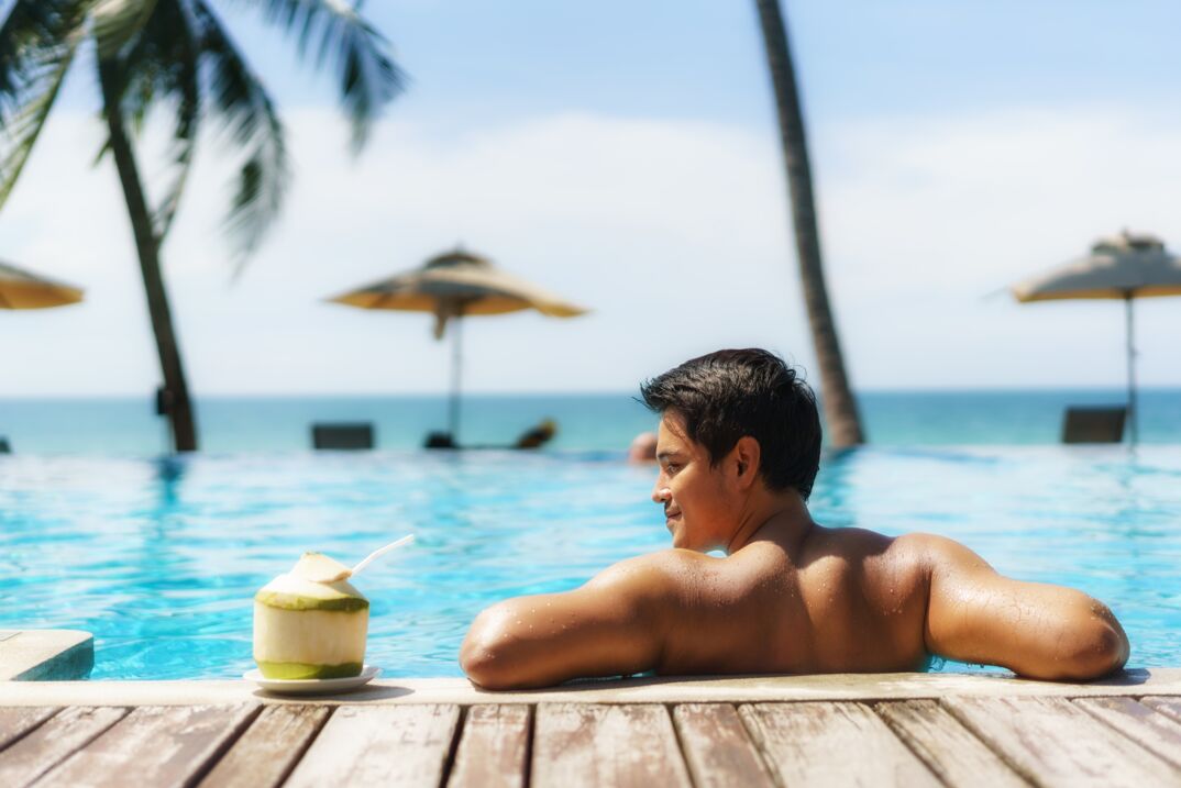 Asian man resting his shoulders by the pool ledge and smirking his face to the side. 